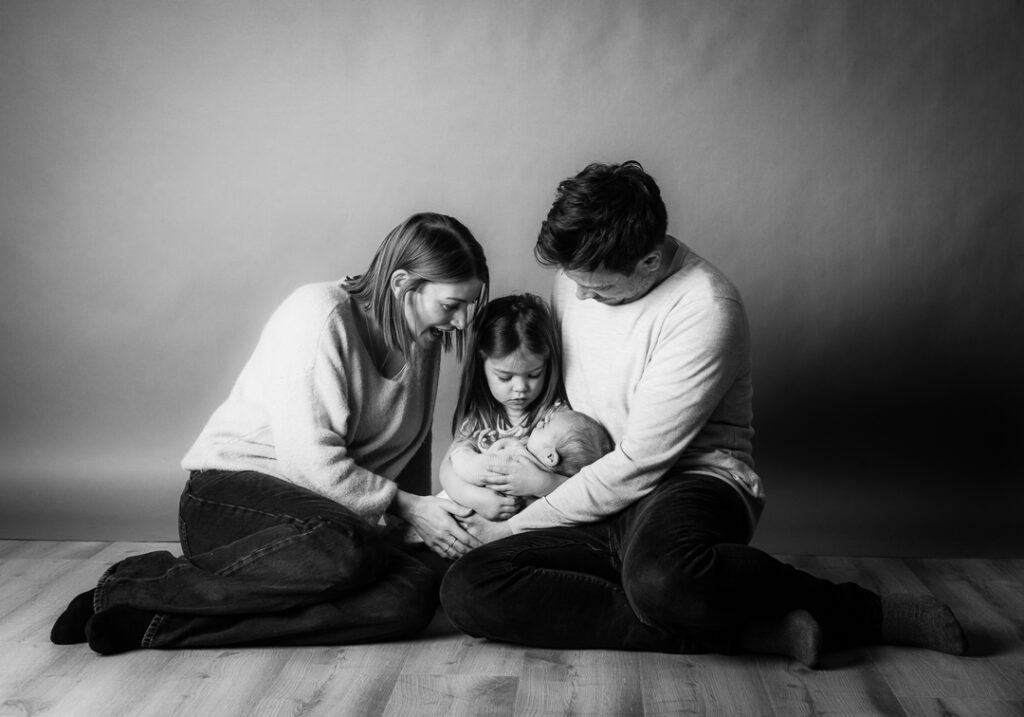 Newborn baby held by sibling and parents during a tender black and white studio portrait at Stables Photography Studio in Monmouthshire
