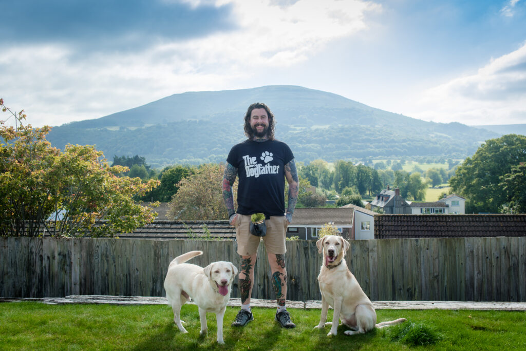 Rich from The Dogfather Abergavenny standing with two Labradors in front of a scenic mountain view, supporting the Stables Photography Studios dog portrait promotion.